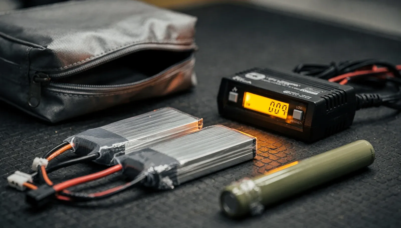 Duas baterias LiPo, um bastão NiMH, um testador digital de células e uma bolsa de carga sobre superfície escura