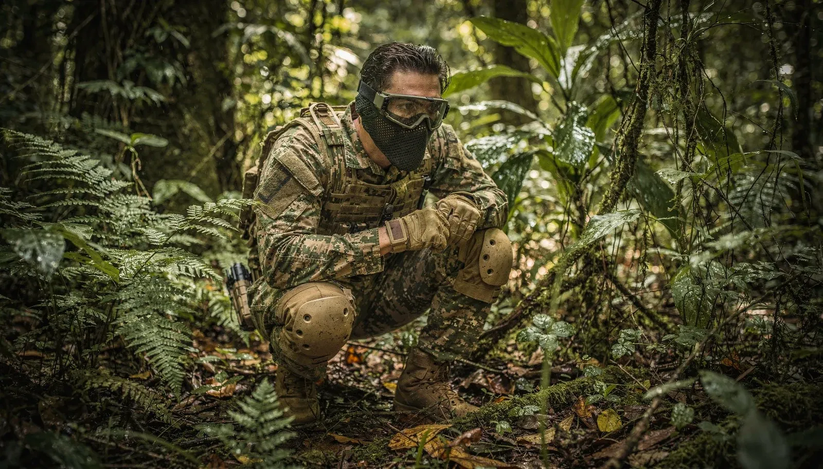 Jogador de airsoft com farda camuflada, máscara e joelheiras agachado no meio da mata fechada