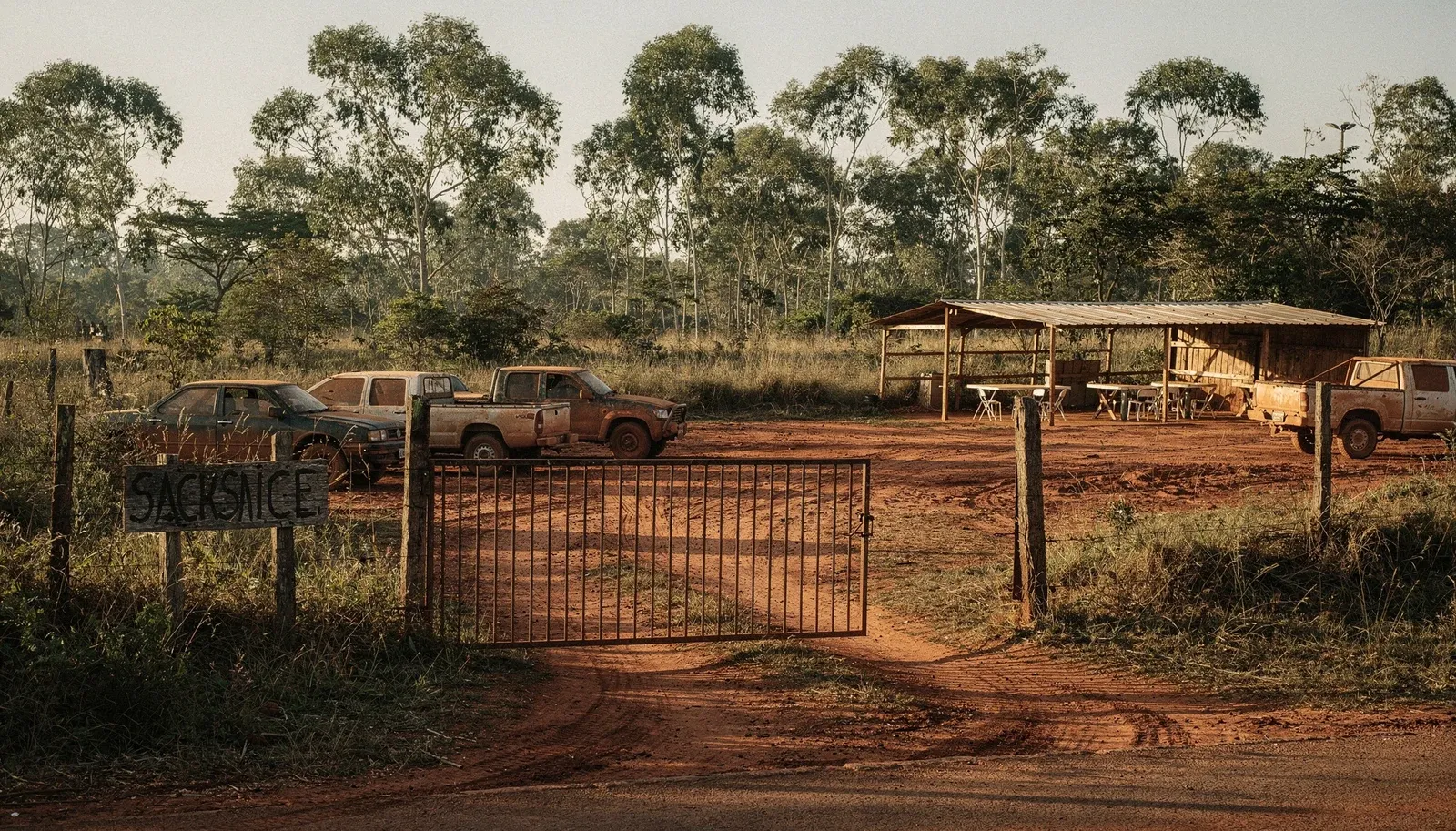 Portão de entrada de um campo de airsoft em estrada de terra, com carros estacionados e tenda da safe zone ao fundo