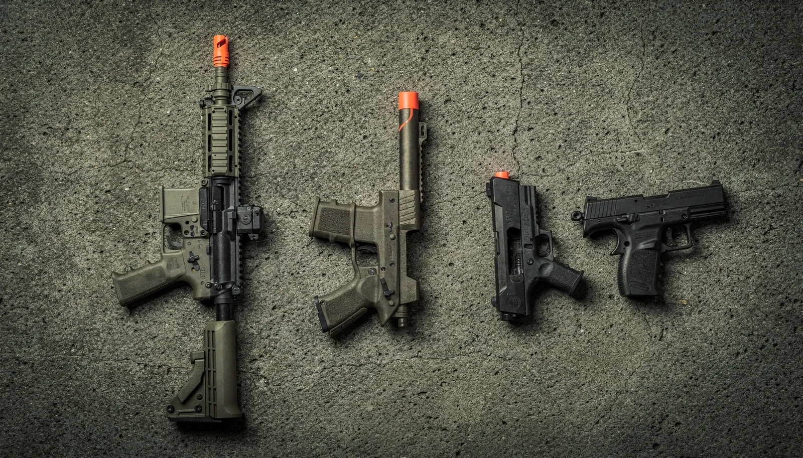 Quatro réplicas de airsoft com ponta laranja alinhadas lado a lado, vistas de cima: fuzil, submetralhadora e duas pistolas