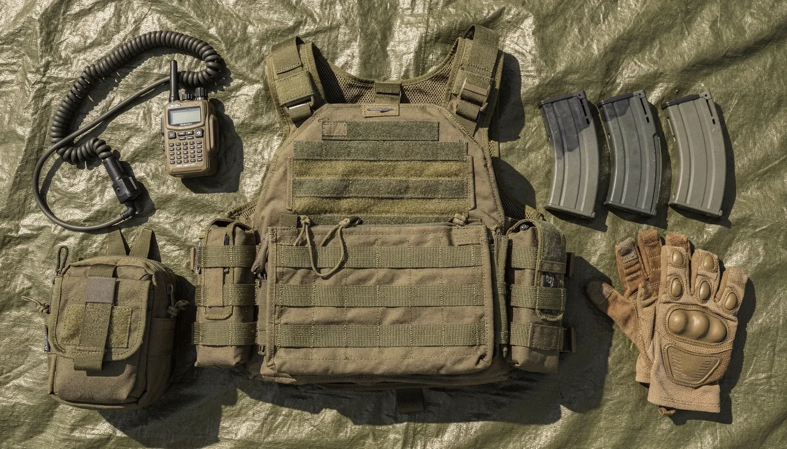 Loadout de airsoft organizado sobre lona: colete tático, rádio, carregadores, pouches e luvas
