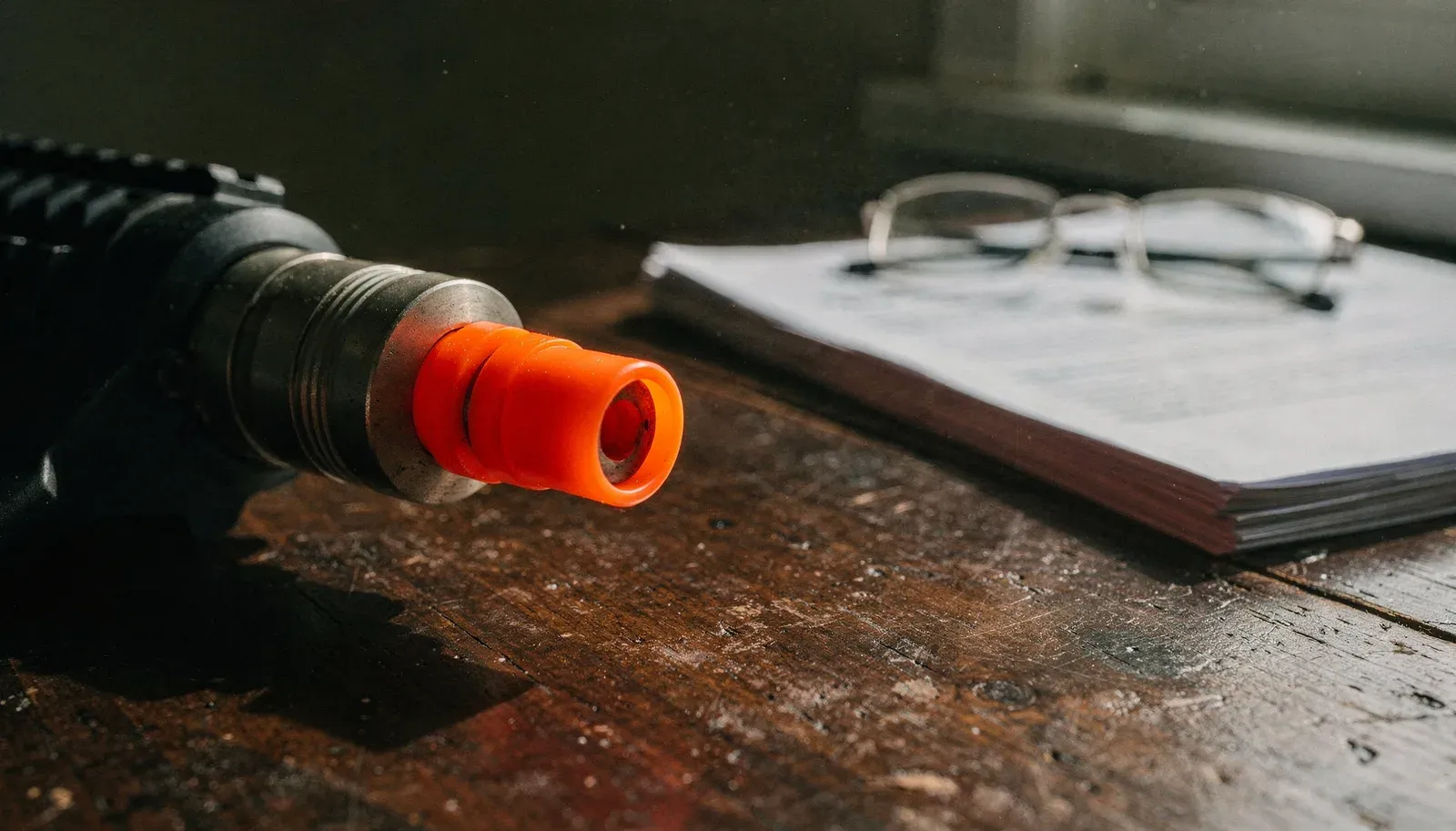 Ponta laranja do cano de uma réplica de airsoft sobre mesa de madeira gasta, com documentos impressos e óculos de leitura desfocados ao fundo