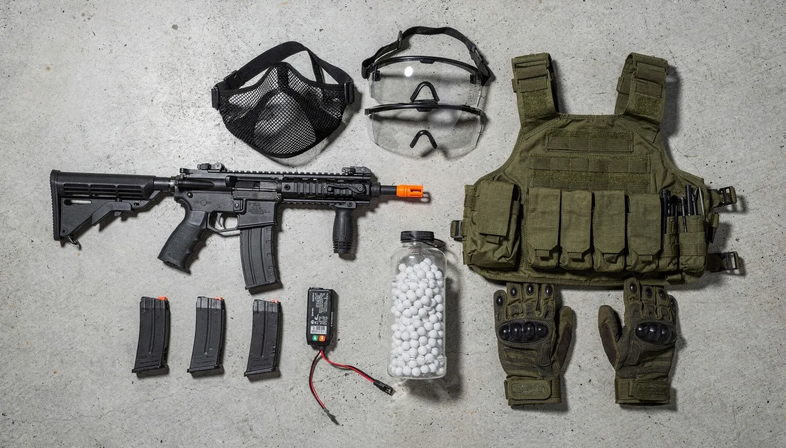 Kit completo de airsoft visto de cima: réplica, máscara, óculos, colete, carregadores, bateria, pote de BBs e luvas